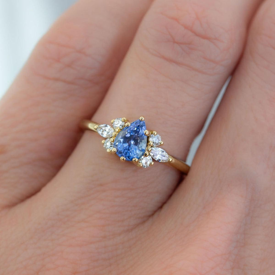 Pear blue sapphire cluster ring with round and marquise shape white diamonds in 18K yellow gold on hand by Amy Jennifer Jewellery